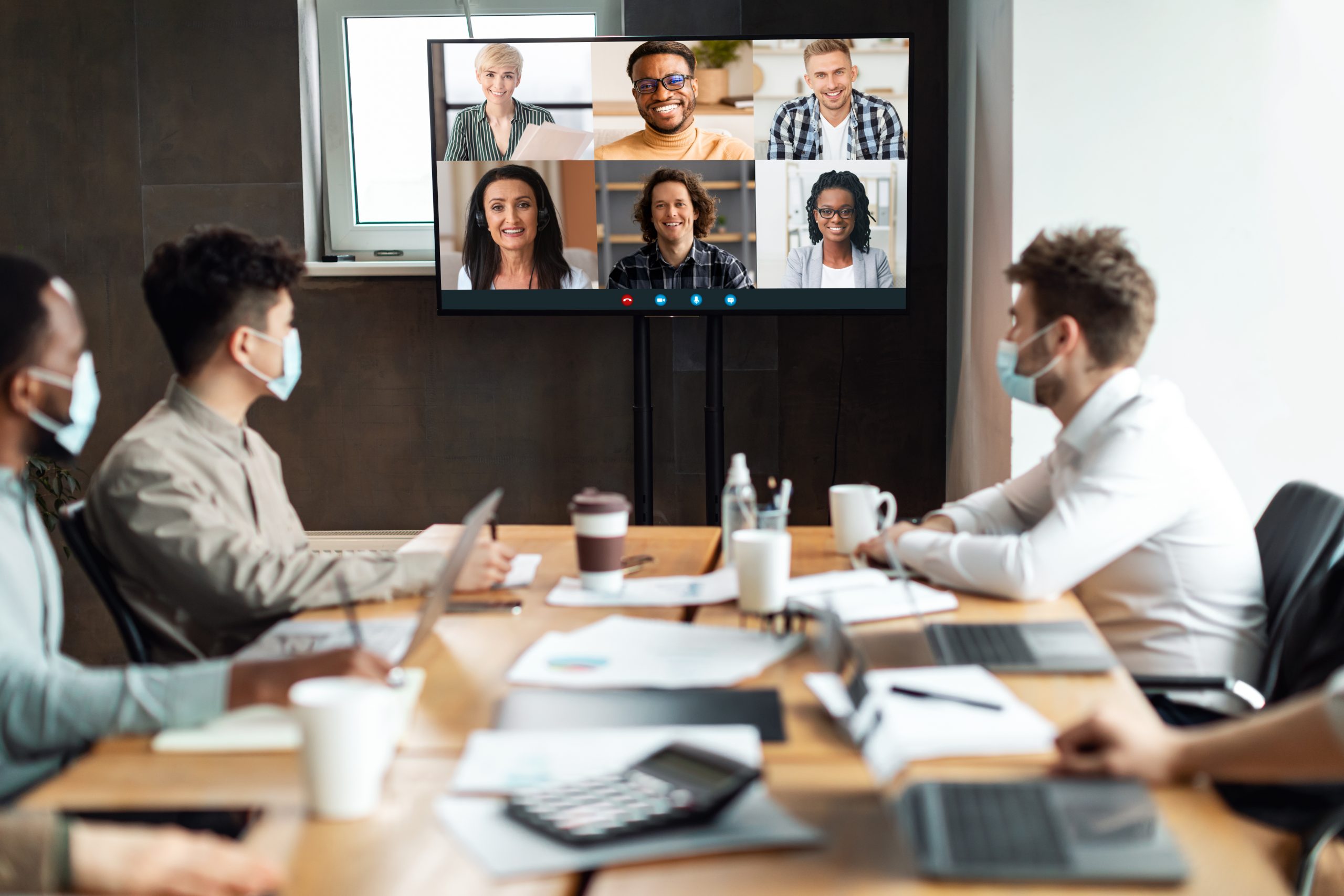 Distant Virtual Meeting Group of diverse business people in masks making online videocall with colleagues, sitting in boardroom, looking at tv screen, talking on web with remote workers