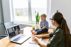 Stay at home. Mother working from home, she talks online with a coworkers via video call on laptop, a cute kid near