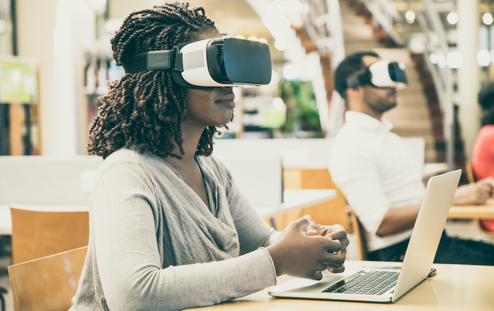 Adult students using VR simulators for work on project in library. Man and woman wearing virtual reality glasses, sitting at desks with laptops, holding air. Augmented reality concept