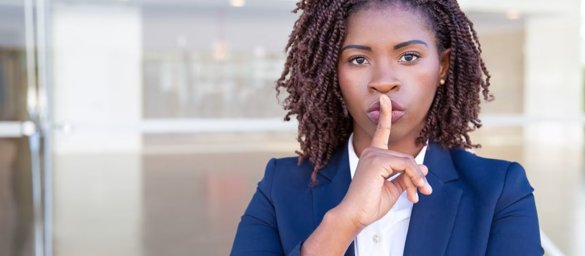 Serious silent professional keeping secret. Young black business woman standing at outdoor glass wall, applying index finger at mouth, making shh gesture. Conspiracy concept