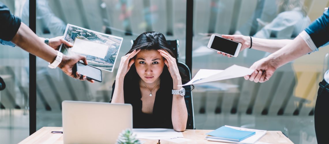 Young disappointed woman feeling headache from deadline surrounded by colleagues at workplace, multi-purpose female employee,tired of work and exhausted, stress in office. Overworked female in confuse