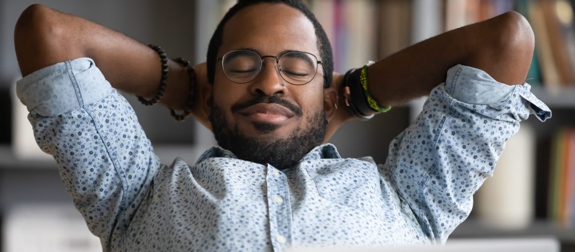Close up calm African American businessman relaxing daydreaming at workplace, satisfied young man sitting with hands behind head, leaning back in comfortable office chair with closed eyes