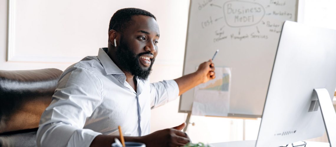 Handsome confident influential african american business coach with beard conducts online training for colleagues or students sitting at table and pointing at flipchart while look at computer screen