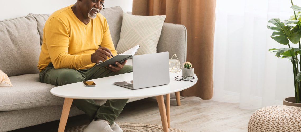 Senior African American Man Using Laptop And Taking Notes Sitting On Couch At Home, Side View. Mature Male Freelancer Working Online Wearing Headphones Indoors. Freelance Career Concept