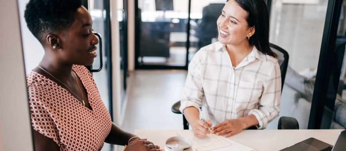hiring and recruitment methods based on skills based hiring when considering job candidate CVs and resumes where two ladies are chatting at a table in a work environment
